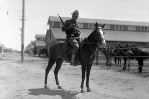 A mounted cavalry soldier poses while pouring tobacco into his rolling paper, outside a row of stables, Fort Logan, Colorado. He wears a sweater, jodhpurs, gaiters and has a rifle slung over his shoulder.