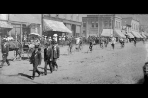 Men march and ride bicycles in a parade in downtown Denver, Colorado. Women ride in a horse-drawn wagon as part of the procession. Signs on buildings read: "Koehler's," "Crooke & Fuller Mine & Mill Supplies," and "Sam Wittow."