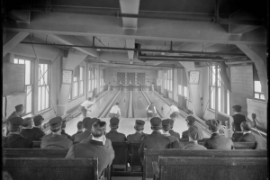 Interior view of a bowling alley in Denver, Colorado; shows lanes, scorekeepers lettered: "Brunswick-Balke-Collender Co.," and rows of seated men wearing Denver Tramway Company conductor's hats.