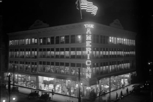 Nighttime rooftop view of the American Furniture Company store at 16th (Sixteenth) and Lawrence Streets in downtown Denver, Colorado. The four-story building has an electric sign in the shape of a U. S. flag. Automobiles and pedestrians are nearby.