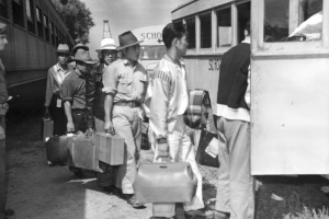 Japanese men arrive from the Merced Assembly Center, California, by train, carrying suitcases and boarding school buses on their way to Camp Amache, Granada Relocation Center, Prowers County, southeastern Colorado. A United States soldier in uniform stands next to the passenger train car and looks on as the line of evacuees board the bus.