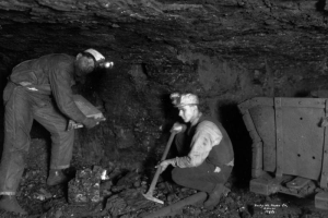 View of two miners working in an underground coal mine, Lafayette, Colorado. One man squats and uses his pickaxe on a chunk of coal, the other holds an unidentified piece of equipment. An empty ore cart is next to them.