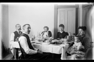 Two men, two women, a girl, and a baby are seated for a meal, probably in Denver, Colorado. Items on the table include: a tablecloth, plates, cups, a coffee pot, a water pitcher, glasses, silverware, and food.