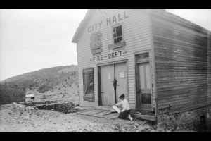 A woman (Muriel Wolle?) is sitting at the entryway of the City Hall and Fire Department building in Nevadaville, Colorado. The name of the building is painted on the facade, and surrounds two broken and boarded up window bays. The woman is holding a sketchpad in her hands.