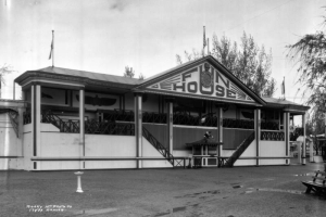 Exterior view of the Fun House at Lakeside Amusement Park, Lakeside (Jefferson County), Colorado. The building has French colonial features including paired exterior staircases, a pavilion roof and thin wooden colonettes. Art Deco designs decorate the columns and the sign reading "Fun House." A face mask and smiling clock are located on the gable. A clown head is attached to the porch, and a monkey figure sits on top of the ticket booth.