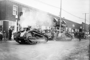 Two military tanks parade on a Arvada street, Jefferson County, Colorado. The lead tank's cannon smokes; drivers can be seen through small  front hatch doors. Arvada Chamber of Commerce sponsors the parade during the third Annual Harvest Festival. Spectators look on from the sidewalk and automobiles are parked curbside.
