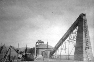 A telescope larger than the Brandon schoolhouse has a large tower with ladders attached to the side of it and a tube that forms an angle with the schoolhouse. Another mechanical device is attached to the one- story schoolhouse.