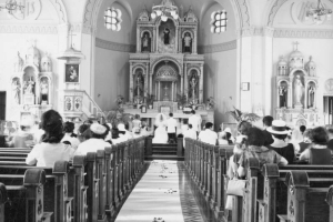 Interior view of the wedding of Lupe Morales and Gene Vigil at St. Cajetan's Catholic Church, 1190 Ninth (9th) Street, in the Auraria neighborhood, Denver, Colorado. The couple kneel in front of the altar, shows religious paintings and statues. People are in the pews, and rose petals are sprinkled along the aisle.
