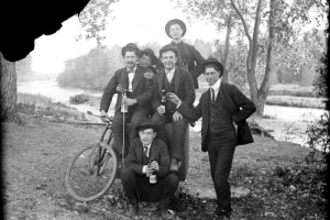 Outdoor portrait of men with a bicycle and bottles of beer possibly near the South Platte River in Denver, Colorado. They wear suits, bowties, and wide-brimmed hats.