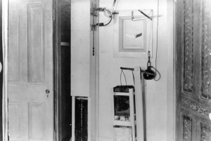 Levers, switches, and a pulley with a weight and a square can are part of the automatic hanging machine at the State Penitentiary in Canon City, Colorado. Wood molding frames a slotted window in the wall, and doors flank the apparatus. 