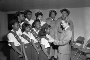 A group of African American (Black) girls and women in Girl Scout uniforms stand in formation to receive badges. A white woman in a dress uniform pins a badge reading: "Be Prepared," on a young girls sash. Other badges indicate that the girls belong to Troop 657, Metro Denver.