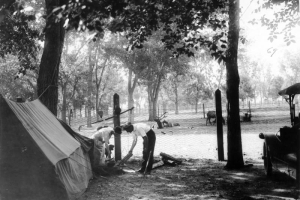 A man and woman arrange logs for a fire at the City Park automobile (auto) camp for tourists in City Park, Denver, Colorado. The woman holds a pot lid and prepares to cook.  Pots and pans hang from a fence post and a canvas tent is set up nearby. Elk lay in a fenced enclosure in the distance.