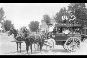 A group of people poses in the "Delta House Smith Bro's. Livery and Transfer" wagon, parked in the middle of unpaved Main Street in the town of Delta, Colorado. Men, women, and children sit in the cab, and two people sit on it. A boy sits next to the driver, who holds reins and a whip. The harness of one of the horses reads "Smith Bros. Livery."  Trees and commercial storefronts are in the background.