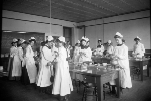 Girls in smocks and caps stir food and peel apples in a cooking class at Mitchell School in Denver, Colorado. Shows a coffered ceiling, long wooden tables with burners, and recipes on the blackboard.