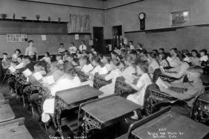 Students sit at desks in a classroom in Denver, Colorado. Their books are open and a few students stand at the front of the classroom. Reproductions of classical Greek sculptures are mounted on shelves. Writing on the chalkboard reads: "Grade 8A, Building 42, June 1919."