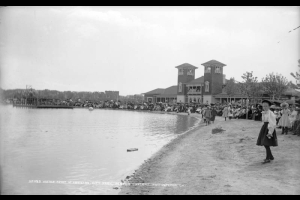 A crowd of people gather around the pavilion and line the shores of City Park Lake (Ferril Lake) in City Park, Denver, Colorado. Men and women are on the balcony of the pavilion. A rowboat and a bandstand are in the lake.