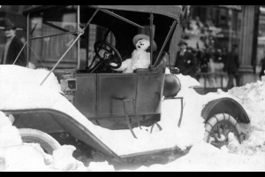 A snowman wears a hat as it sits in the driver's seat of probably a Model T Ford after the snowstorm of 1913 in Denver, Colorado. Pedestrians walk down sidewalk near piles of snow.