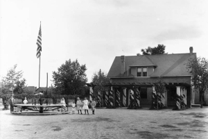 Children pose next to the merry-go-round and flagpole at the Elyria Community Center in Elyria Park, 4809 High Street in the Elyria Swansea neighborhood of Denver, Colorado. Shows a two story brick building with a dormer and a stone column pergola.