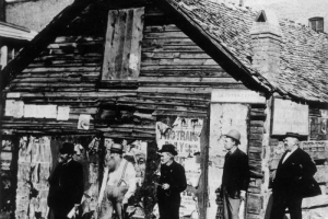 Five men stand in front of the Rocky Mountain News building near 1301 Walnut Street in the Auraria neighborhood of Denver, Colorado. Shows a single story wood frame building with brick chimney. Four men wear dark suits and straw boaters or bowlers. One man wears suspenders and leans on a cane. A Midland Railroad advertisment is posted on the side of the building.