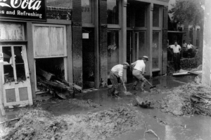 Men use shovels to clear mud from the Cherry Creek flood after the Castlewood Canyon Dam break in Denver, Colorado. Teenage boys look on; storefront signs read: "Coca Cola," "Patterson's Eggs," and "233."