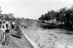 View of a Cherry Creek flood in Denver, Colorado after the Castlewood Canyon Dam break; shows torrents of muddy water in standing waves. People look on from the side; boys wear plastic visors.