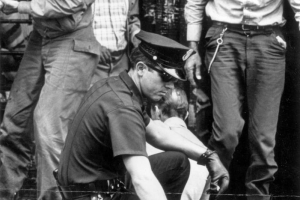 A policeman squats by the side of Larimer Street in the central business district of Denver, Colorado and dumps a bottle of liquor onto the pavement. The police officer wears a short sleeved summer uniform, sun glasses, and leather gloves. A walkie-talkie is in his pocket and keys dangle from his belt. Men on the sidewalk watch the policeman.
