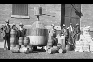 A group of men wearing suits and hats stand near a large still and barrels of liquor near Greeley (Weld County), Colorado. One man leans his arm on a pile of sacks with labels reading: "100 lbs, Cerelose, Product Refining Co., New York, U.S.A."