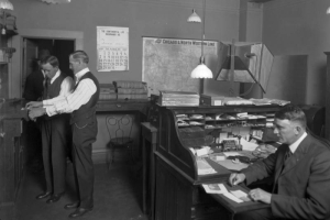 A clerk presses the inked finger of a man onto a paper at the Denver Police Department offices in the City Hall Building, located at Fourteenth (14th) Street and Larimer Street in Denver, Colorado. Another man sits at a roll top desk near a photographic lamp and backdrop.  A map labeled "Chicago & North Western Line" hangs on the wall. A calendar advertising "The Continental Life Insurance Co." turned to March 1929 hangs nearby. Light fixtures hang from the ceiling.
