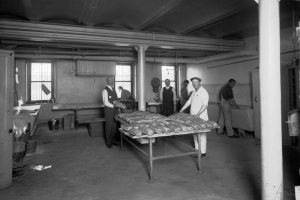 An inmate stands near a table covered with bread loaves at the Municipal Bakery in the County Jail, Denver, Colorado. Other inmates work at sinks and tables forming loaves. Denver City Councilman, John Conolon holds a loaf of bread in his hand, and Warden Frank Kratke stands near a clock.