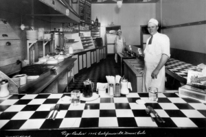 Interior view of the Pig Parlor restaurant in downtown Denver, Colorado; men pose in the kitchen behind a checkered counter and condiments. Decor includes a deco ceiling lamp, a mural of pigs, a grill, coffee machines, and a menu.