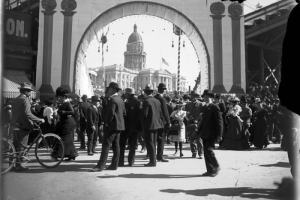 People walk in a baseball stadium (probably Broadway Athletic Park) at Colfax Avenue and Broadway in Denver, Colorado, during the Festival of Mountain and Plain. The Colorado State Capitol building shows through the entry arch.
