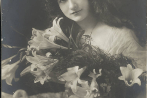 Maude Fealy poses for a studio bust portrait, possibly for the role of "Filiberta" in "The Cardinal." She has long, wavy hair; wears a bracelet, a pearl necklace, and a dress with gathered, sheer fabric and beadwork; and holds a bouquet of lilies.
