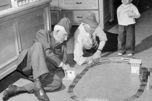 Interior view of two old men as they play with a "Union Pacific" railroad set, a boy beside them cries. The men wear railroad caps. The model bridge reads: "Laxkawanna."