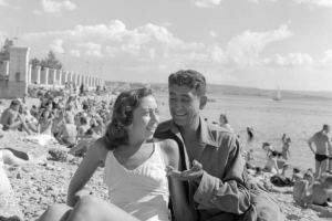 A Tenth Mountain Division soldier enjoys the beach at Trieste with a woman. Both wear bathing costumes and pose for the camera. Behind them the beach is filled with other sun worshipers.