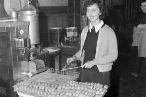 Red Cross doughnut girl, Deborah Bankart, poses standing by an automatic doughnut making machine with one full and one partially-filled tray of doughnuts before her, which she handles with two sticks. She wears a sweater vest over a shirt and a cardigan and a Red Cross pin. The trays and the machine are set on packing crates. Bankart was the first doughnut girl with the Tenth Mountain Division in Italy.