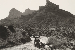 View of wagons, one with a covered wagon frame, pulled by teams of six horses on a dirt road in Arizona. Shows men and supplies in the wagons, rocky buttes, brush and saguaro cacti near the road on the Apache Trail in probably the Superstition Mountains.