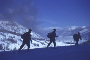 Three Tenth Mountain Division soldiers cross-country ski above Camp Hale. They appear in silouette in the foreground of the image with the Pando Valley and Camp Hale, Colorado, brightly lit by the sun, in the background. Visible in the air above the valley is the coal smoke from passing freight trains.