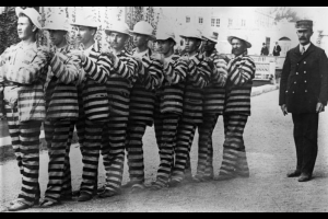 Convicts at the State Penitentiary in Canon City, Colorado, wear uniforms marked with wide, horizontal stripes. They stand in a line, each with his hands on the shoulders of the man in front of him. A uniformed guard in a double breasted jacket and conductor's hat looks on.