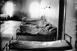 Black and white convicts lie in cots in the hospital at the State Penitentiary in Canon City, Colorado. The walls are partly tiled; electric lights and a steam radiator furnish the interior.