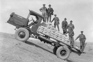 View of a Coleman flatbed truck on an incline with the city of Denver, Colorado, in the distance. A group of well-dressed men and workers ride on the truck loaded with sacks of probably sand. The surrounding landscape is barren. The state capitol building is visible in the distance.