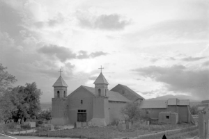 The Spanish Colonial mission at Santa Cruz, New Mexico is adobe and has two pitched-roof steeples with crosses on top. A woman with a dark shawl wrapped over her head and shoulders walks in the foreground down a path at the side of the mission.
