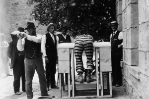 Bent over a wooden, upholstered frame, a prisoner at the State Penitentiary in Canon City, Colorado, wears patched, striped prison pants. His ankles are shackled, and a man in a vest, gold chain and cowboy hat swings a hinged paddle, targeting the convict's buttocks. A wooden bucket is on the ground, for wetting the implement. Men in vests, ties, and straw hats look on.