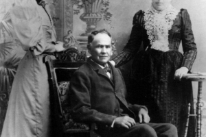 Studio portrait of Magdalena Córdova, daughter of Juan Bautista Córdova, and his wife, María Quirina Sánches. Magdalena wears a long dress with leg of mutton sleeves, and velvet trim. Her hair is curled and pulled back. She stands beside her father who is seated in a chair; he  wears a suit and bow tie, his hair is receding and he is clean shaven. His wife, María wears a dark silk dress with slightly puffed sleeves and a crocheted bertha collar.