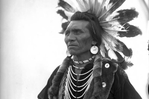 Chief Eagle, a Native American man on the Flathead Indian Reservation in western Montana, poses wearing a warrior's headdress in front of a teepee on the reservation. He wears large, round earrings, numerous beaded necklaces hanging around his neck, an otter sash across his chest, and holds a long spear, possibly a coup-stick, also wrapped in fur.