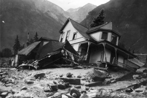 Exterior view of Jack Hawkins residence, on Oak Street, filled with mud and debris from the disastrous Cornet Creek flood on July 27, 1914, Telluride, Colorado; shows two-story wood frame Victorian with front porch and second story bay window tilting on side from flood waters, smaller destroyed structure, and the San Juans in background.