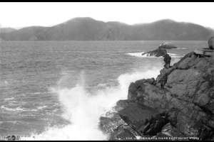 Men stand on rocks as waves splash against the seawall at Fort Point at the Golden Gate channel in San Francisco Bay, San Francisco County, California. Shows the corner of a pillbox.