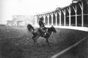 A cowboy rides a bucking horse in a dirt arena during a performance of Buffalo Bill's Wild West Show at Earl's Court in England. The grandstand curves in the right background and is supported by large wood beams.