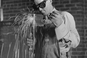 A student welds a piece of metal in a classroom in Greeley, Colorado in Weld County.  He holds the welder in his right hand and the piece of metal in a pair of tongs in his left hand.  He wears a cap, goggles, and heavy work gloves.  He dressed in overalls and a long-sleeved shirt.