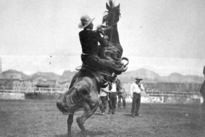 Antonio Esquivel, a champion Mexican vaquero and star of Buffalo Bill's Wild West Show, rides a bucking horse in a dirt arena during a performance of the show at Earl's Court in England. He wears a cowboy hat and a short, dark jacket. A group of cowboys stands in the background watching Esquivel ride. A backdrop with a landscape scene painted on it is at the far end of the arena.