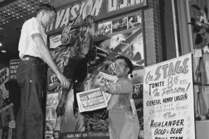 A dwarf hands flyers to a man at the Tabor Theater, 1014 16th (Sixteenth) Street in Denver, Colorado. Signs read: "Invasion U.S.A.," "Prince of Pirates," "A Bomb Blasts in U.S. Cities," and "On Stage Tonite 8:15 p.m. in Person! General Henry Larsen, Chief Civilian Defense Plus Highlander Blue and Gold Drill Team, World Famous Tabor."